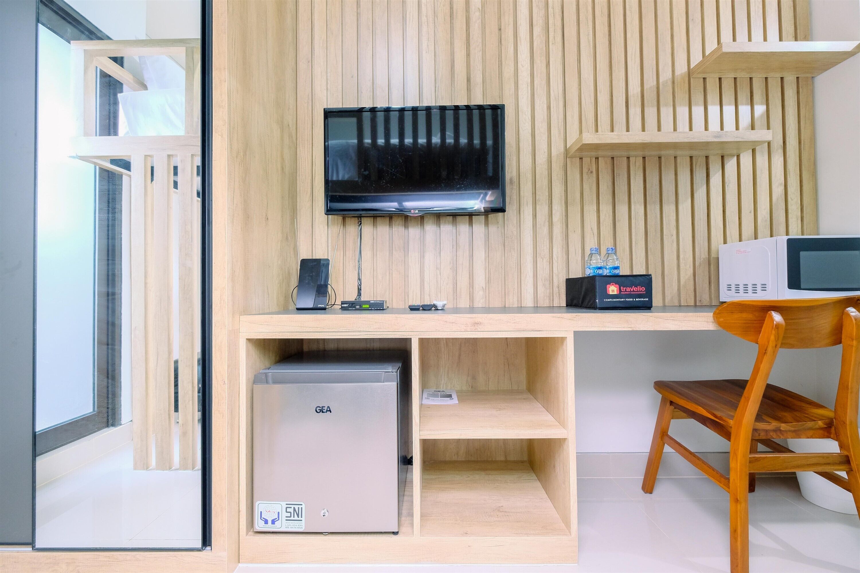 Una habitación moderna con un escritorio de madera, un refrigerador GEA, una televisión, un microondas y una silla de madera.