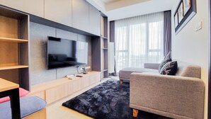 A modern living room with a flat-screen TV mounted on the wall, a grey sofa, a black rug, and wooden shelves. - Scenic And Comfortable 1Br Apartment Branz Bsd City (Pagedangan)