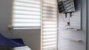 Une chambre avec une fenêtre équipée de stores, un téléviseur à écran plat fixé au mur et un lit recouvert d’une couverture bleue.