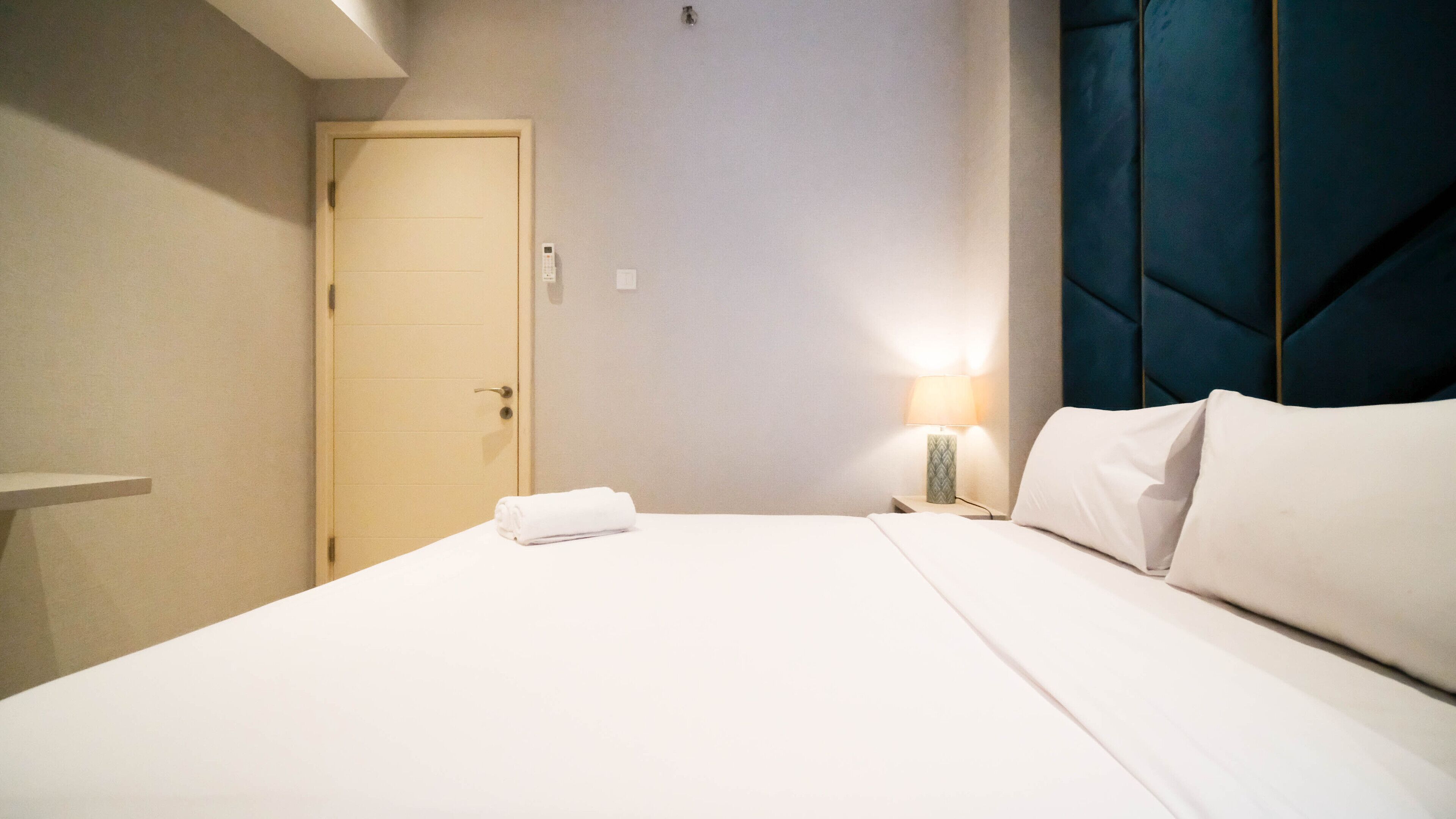 Habitación de hotel con cama, mesita de noche con lámpara, una puerta y cabecera azul.