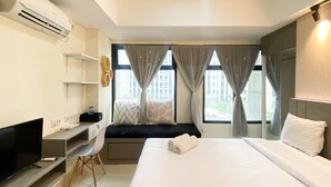 A hotel room with a bed, a desk with a computer, a TV, and a window with curtains. - Cozy Studio Room At Pollux Chadstone Apartment (Cikarang)