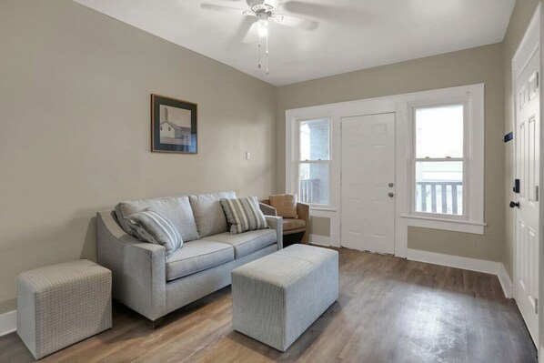 A living room with a grey sofa, two ottomans, a ceiling fan, a door with a window, and a framed picture on the wall. - Cozy 2 Bed - Downtown-West #7 (Indianapolis)