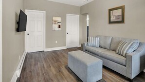 Ein geräumiges Wohnzimmer mit einem grauen Sofa, einem gestreiften Ottomanen und einem Gemälde an der Wand.