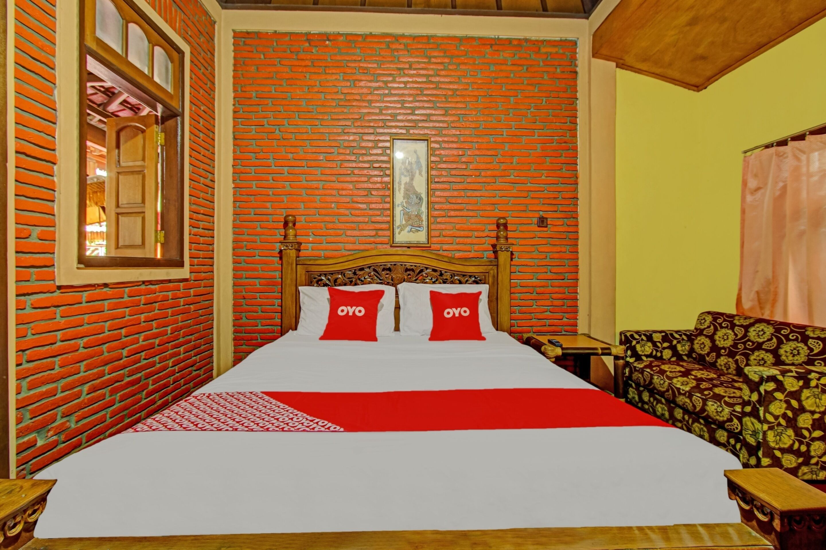 A room with a brick wall, a wooden bed with red pillows, a patterned sofa, and a framed picture on the wall.