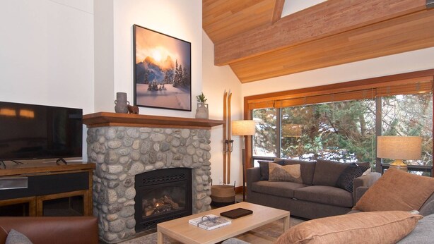A cozy living room with a stone fireplace, a flat-screen TV, a comfortable sofa, and a large window offering a view of trees.