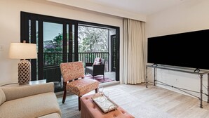 A hotel room with a sofa, armchair, ottoman, a TV on a stand, a lamp, and a balcony with a view of trees and a building.