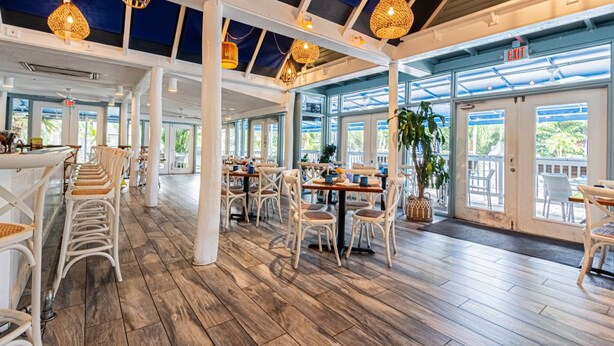 A spacious indoor area with wooden flooring, white chairs, and tables. There are large windows allowing natural light and views of the outdoors.