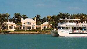 A large, two-story house with a symmetrical facade, a boat named 'DOUBLE SUNSHINE' docked nearby, and palm trees in the background.