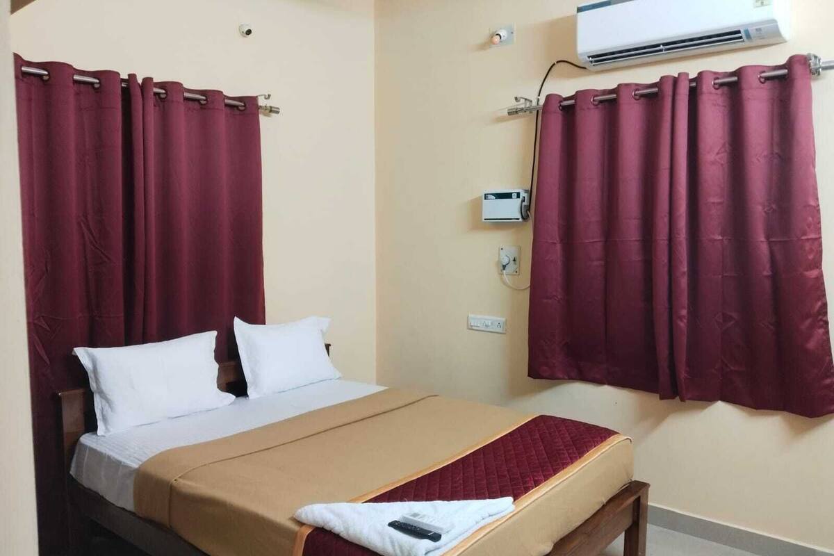 A single bed with a beige mattress, white pillows, and a red blanket. There is a wall-mounted air conditioner and a power outlet on the wall.