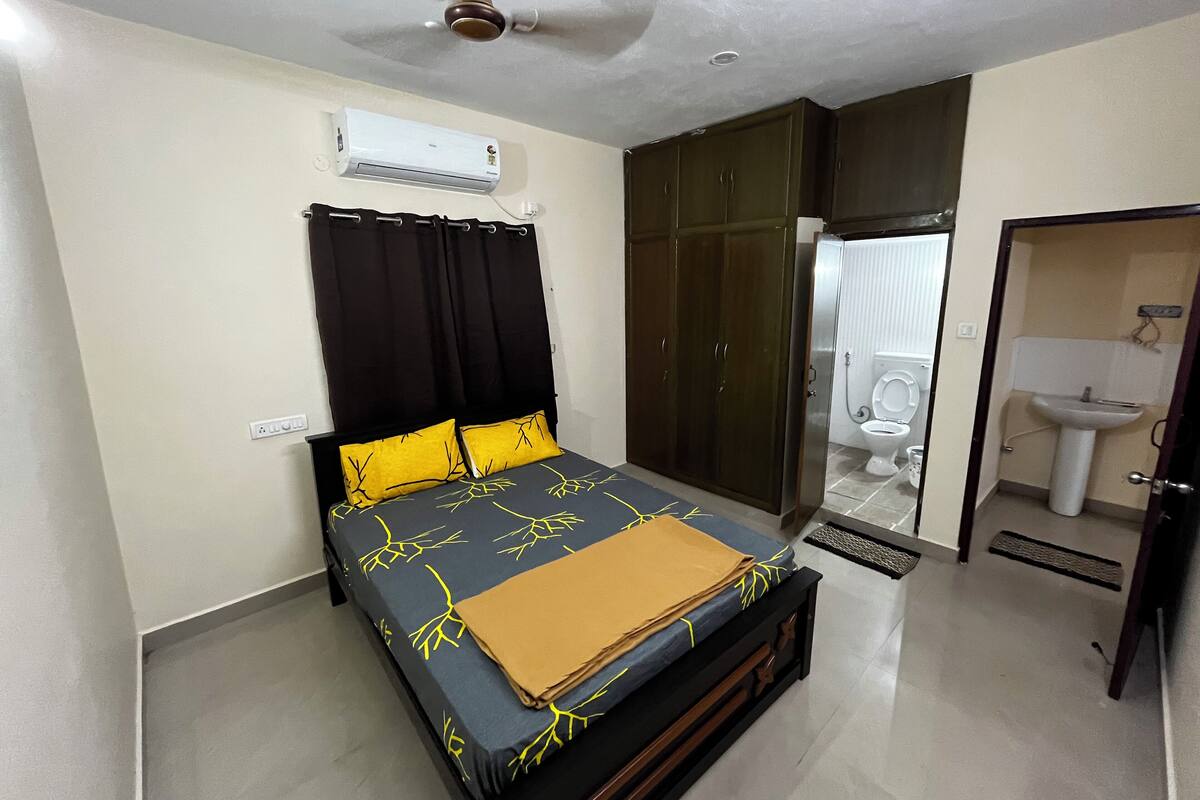 A bedroom with a bed, an air conditioner, a wardrobe, and a bathroom with a toilet and sink.