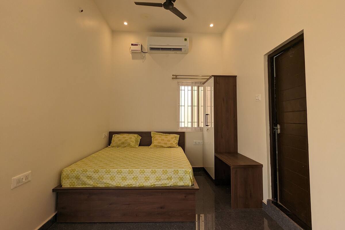 A bedroom with a bed, ceiling fan, air conditioning unit, and a door.