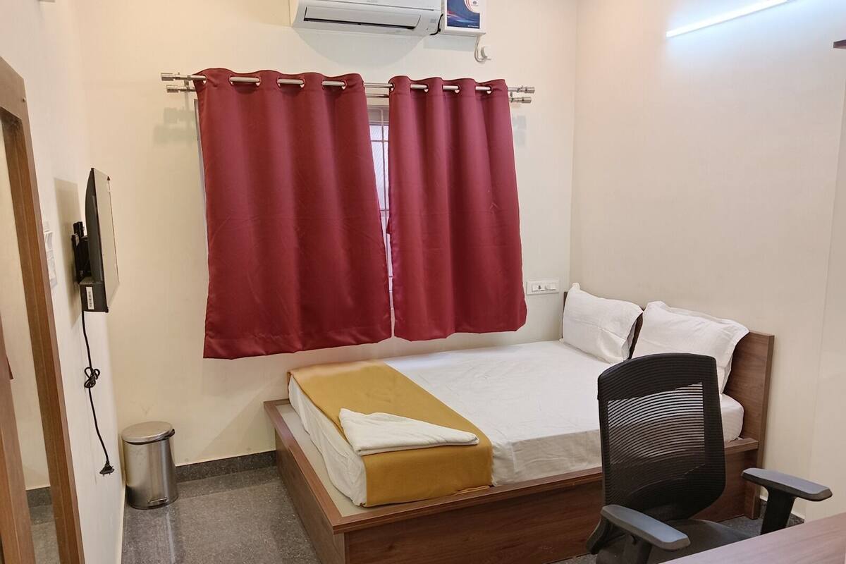 A bedroom with a bed, a chair, a desk, a trash bin, and a curtain.