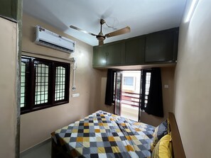 Une chambre comprenant un lit, un climatiseur, un ventilateur de plafond, une fenêtre avec une grille en métal et une porte donnant sur un espace extérieur.