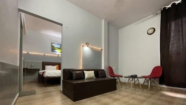 A modern living room with a dark brown sofa, two red chairs, a small table, and a bed in the background. - Abodes Elegant Hills (Dehradun)