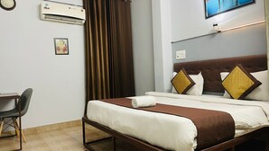 A hotel room with a bed, a desk, an air conditioner, a television, and a window with curtains. - Abodes Elegant Hills (Dehradun)
