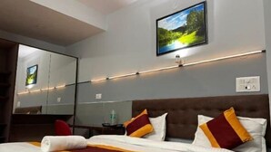 A hotel room with a neatly made bed, a large mirror, a flat-screen TV mounted on the wall, and a view of a green landscape. - Abodes Elegant Hills (Dehradun)