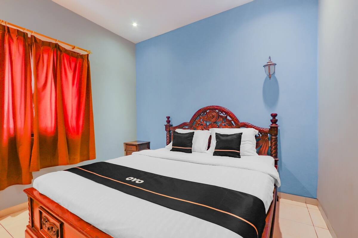 A neatly made bed with a black and white bedspread and a logo, a wooden headboard, a nightstand, and a wall-mounted lamp.
