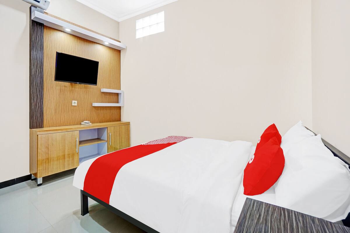 Ein Hotelzimmer mit einem Bett, einem an einer Holzplatte montierten Fernseher, einem kleinen Schreibtisch und einer Klimaanlage.