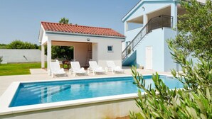 Une piscine avec des chaises longues, un patio couvert et un bâtiment à deux étages surmonté d’un toit en tuiles rouges.
