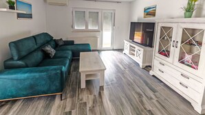 Un salon avec un canapé de couleur turquoise, une table basse, un téléviseur posé sur un meuble et une fenêtre avec des stores.