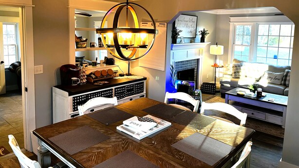 A dining area with a wooden table, chairs, and a chandelier. There is a fireplace in the background and a window with a view. - SC Farmstead Cottage with modern amenities, farm views, and equine visitors (Belton)
