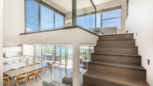 A modern interior with a staircase leading to a second floor, a dining area with wooden chairs and a table, and large windows offering views of the outdoors.
