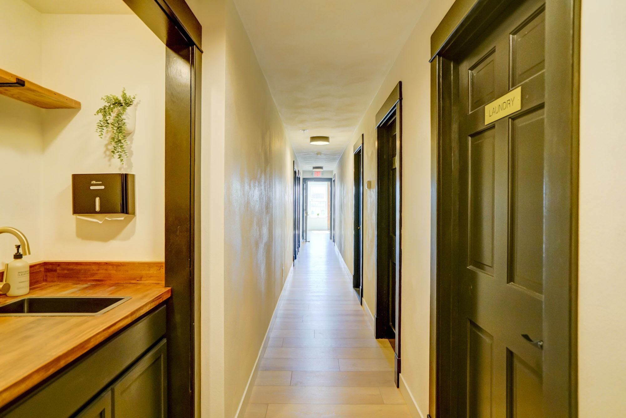 A hotel hallway with a wooden counter, a sink, a plant, a wall-mounted dispenser, and a door labeled "Laundry".