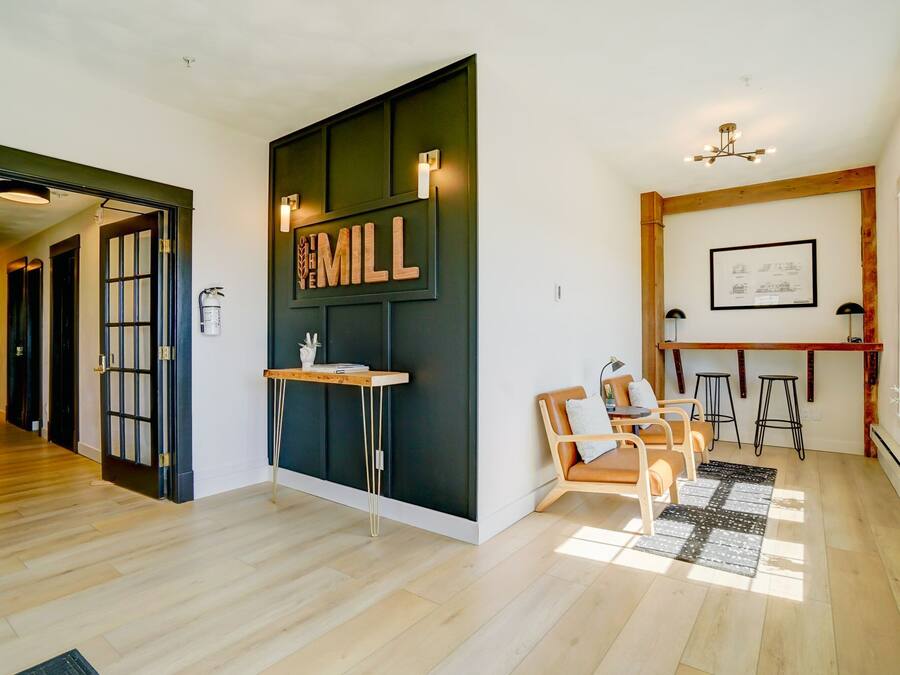 A modern interior with a wooden floor, a dark accent wall with 'THE MILK' signage, a seating area with a table and chairs, and a bar area with stools.