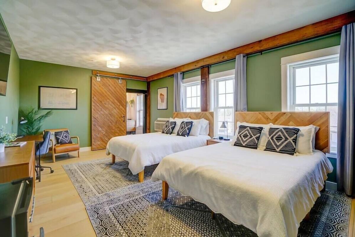 A room with two beds, a desk, a chair, and a large window.