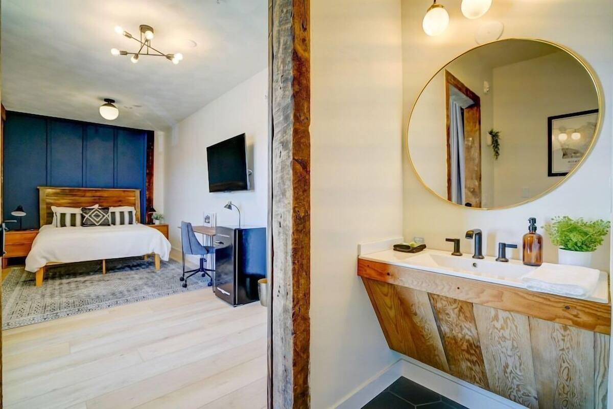 A bedroom with a bed, desk, and chair, a bathroom with a sink and mirror, and a ceiling light fixture.
