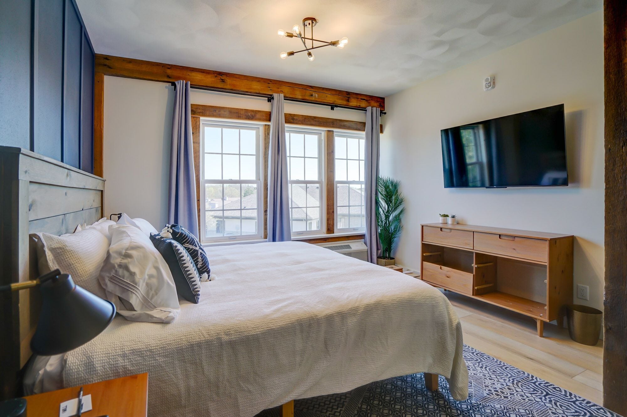 A bedroom with a bed, a TV, a wooden dresser, and large windows.