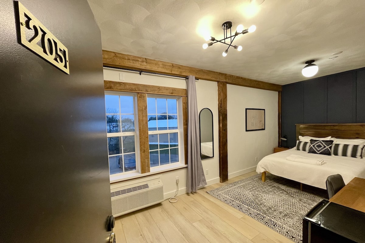 A bedroom with a bed, a desk, a radiator, a window with curtains, and a ceiling light fixture.