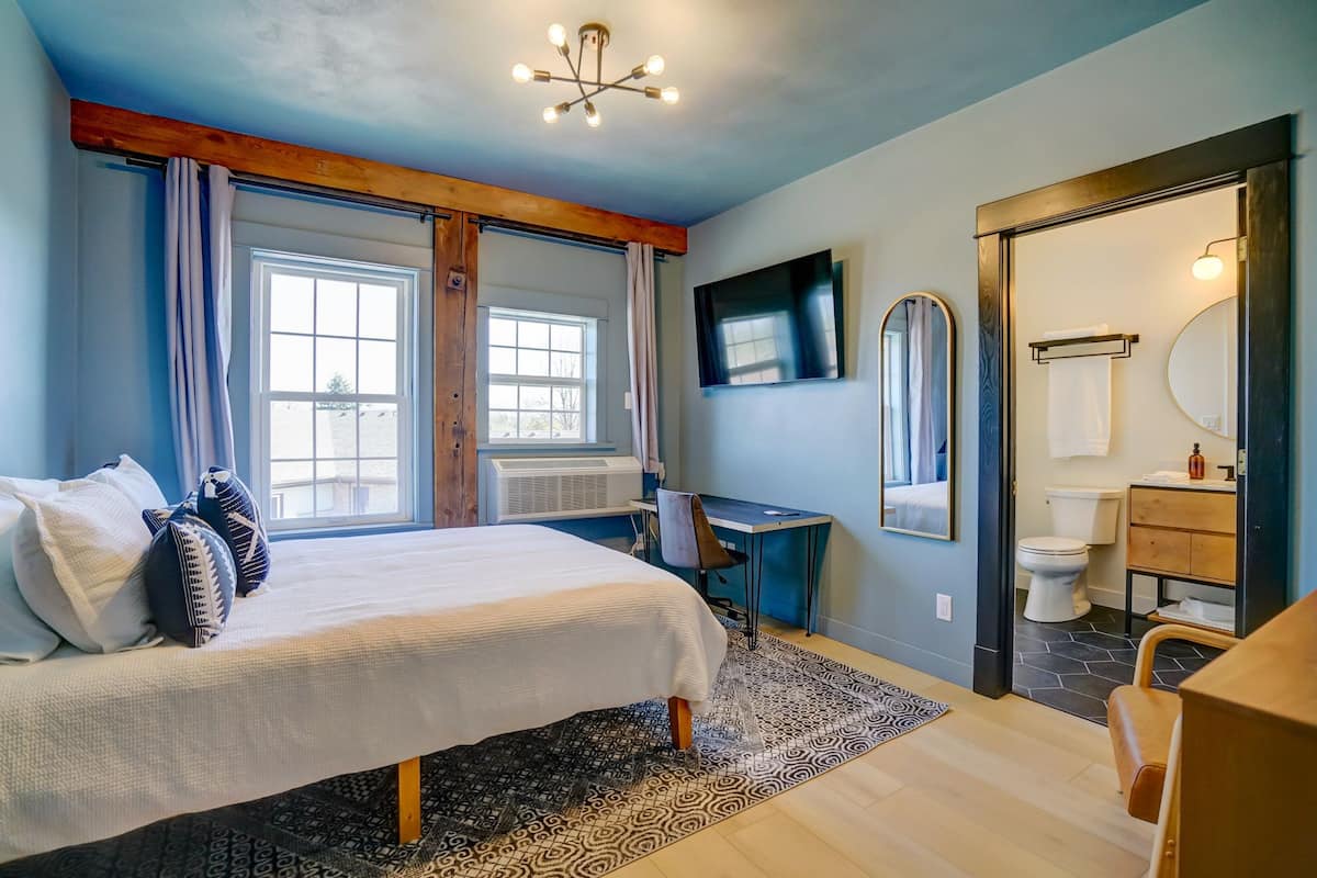 A bedroom with a bed, a desk, a chair, a large window, and a bathroom with a toilet and sink.
