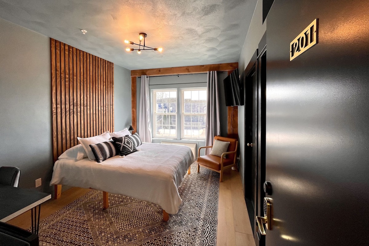 A modern hotel room with a bed, a desk, a chair, a TV, and a large window.