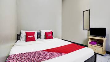 Un lit bien fait, recouvert d’une couvre-lit à motifs rouges et blancs, deux oreillers portant la marque « OYO », un téléviseur posé sur un meuble en bois, et une petite armoire en bois contenant des vêtements pliés.