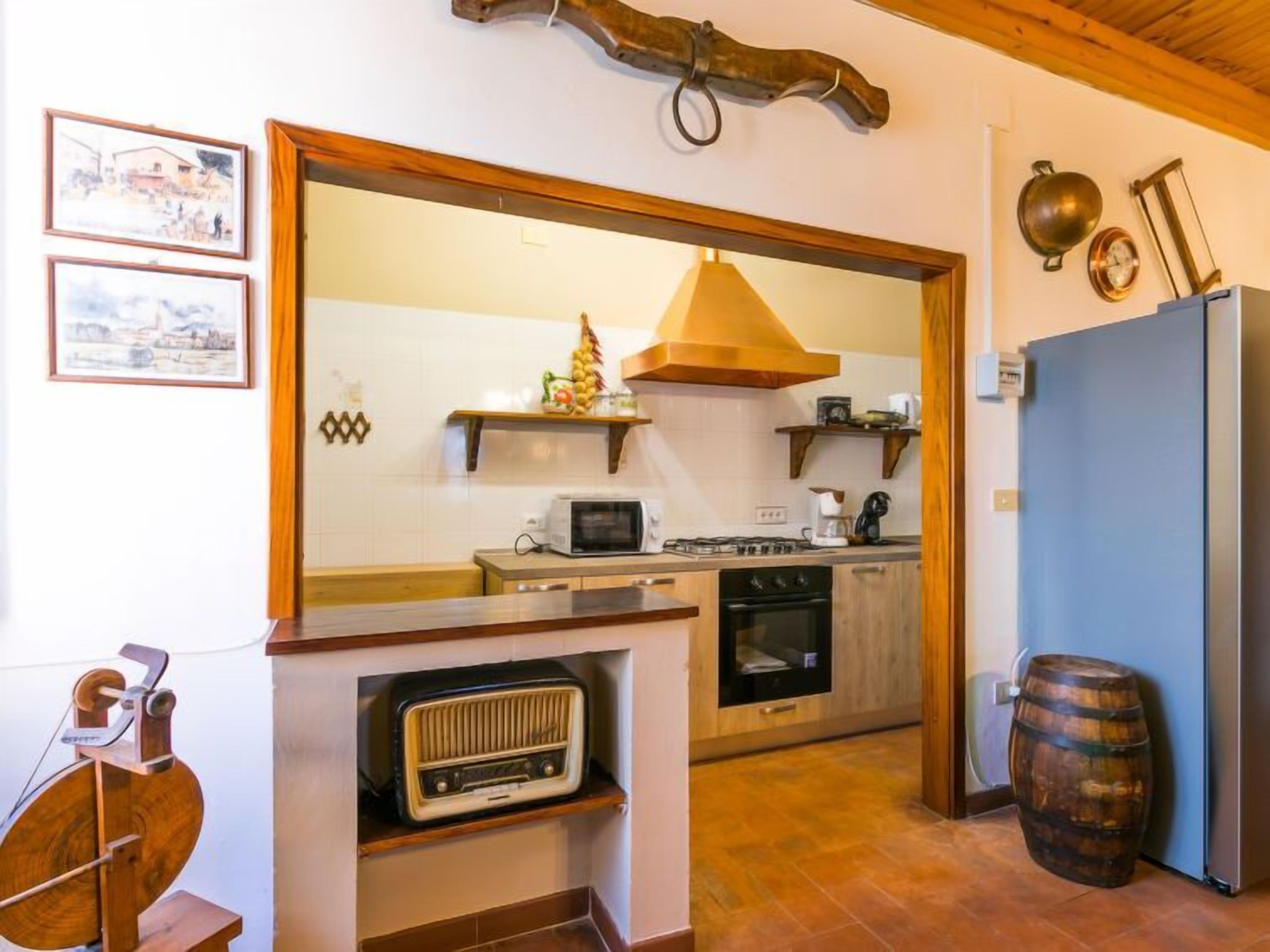 Una cocina con gabinetes de madera, un microondas, una estufa y un refrigerador. Se observa un barril de madera y una radio sobre una estantería.
