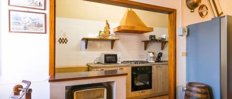 Una cocina con gabinetes de madera, un microondas, una estufa y un refrigerador. Se observa un barril de madera y una radio sobre una estantería.