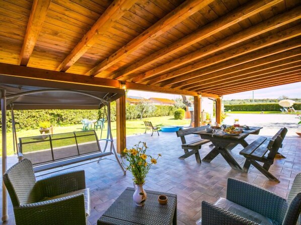 Une terrasse extérieure avec des tables et des chaises en bois, un espace salon couvert et une vue sur un jardin et une piscine.
