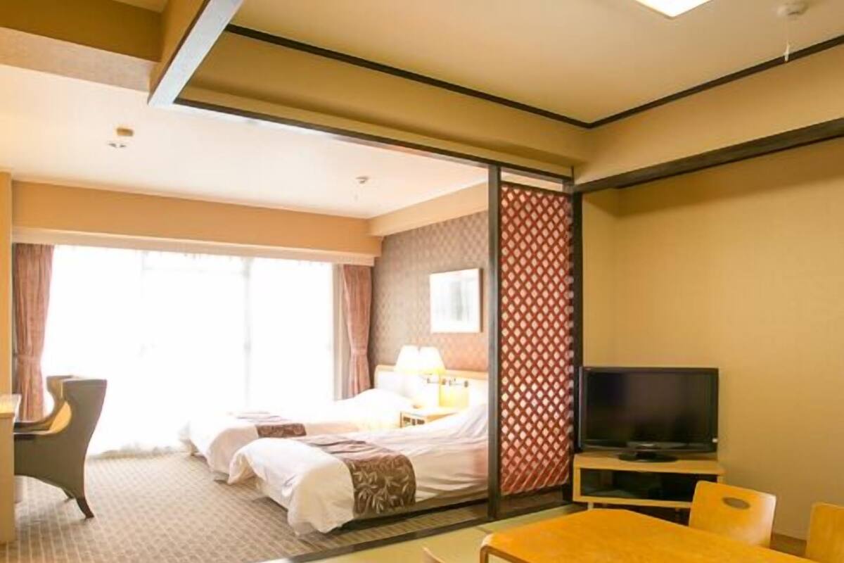 A hotel room with a bed, a TV, a dining table, and a sliding door leading to another room.