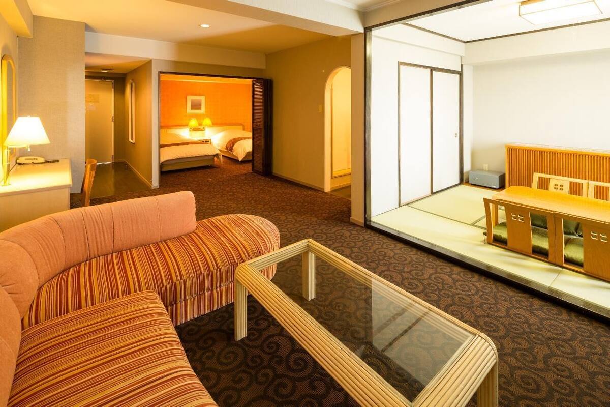 A hotel room with a sofa, a glass-top coffee table, a bed with a headboard, and a small seating area with a bench.