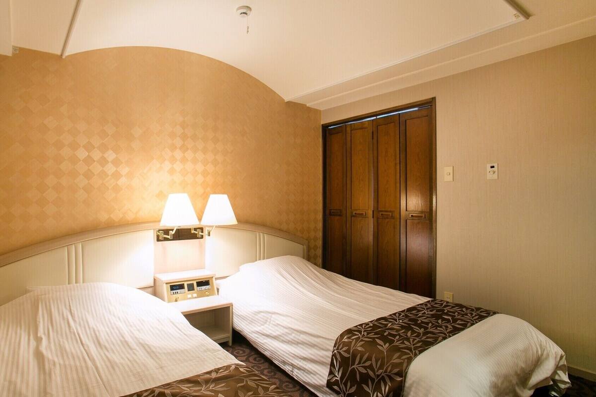 A hotel room with two beds, a nightstand with lamps, a wooden wardrobe, and a ceiling light.
