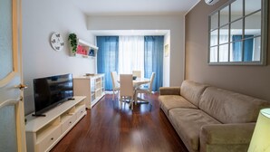 Uma sala moderna com televisão de tela plana, sofá bege, mesa de jantar com cadeiras e área de cozinha com armário branco e uma planta.