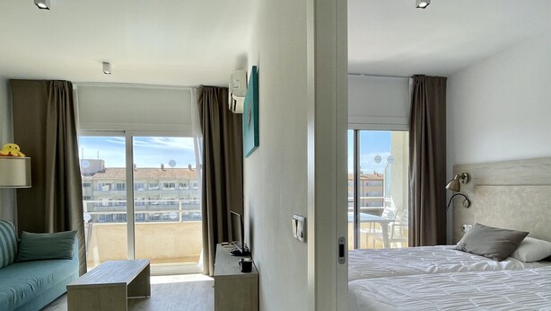 A hotel room with a bed, a sofa, a small table, a TV, and a balcony with a view of buildings. - FESTASTAR 406 (Estartit)