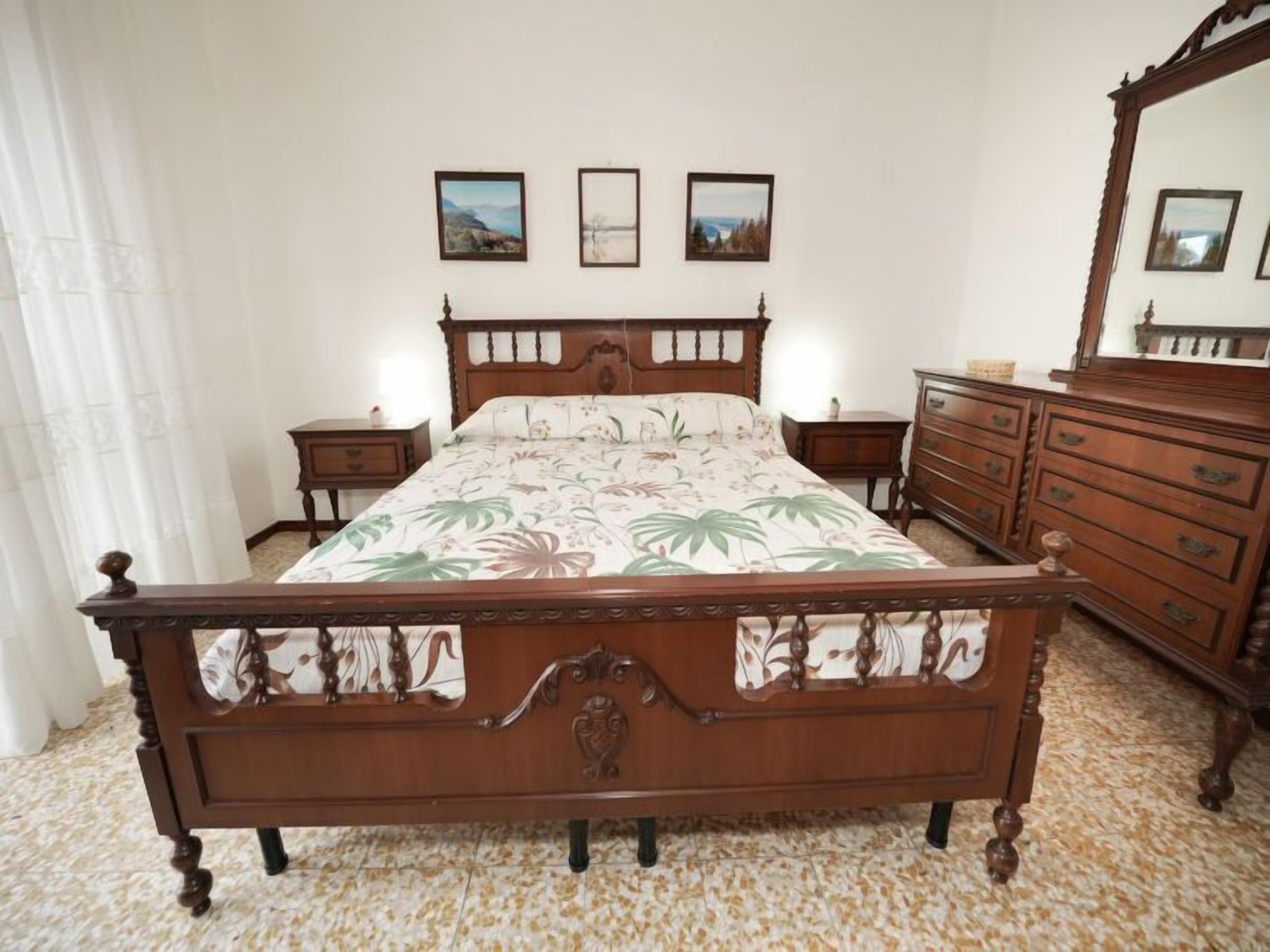 Quarto com cama de madeira, cômoda, mesa de cabeceira e espelho. Há três quadros na parede.