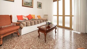 Ein Wohnzimmer mit Sofa, Couchtisch und einem Bett mit gestreifter Decke. An der Wand hängen zwei Bilder und ein Fenster mit Vorhängen.