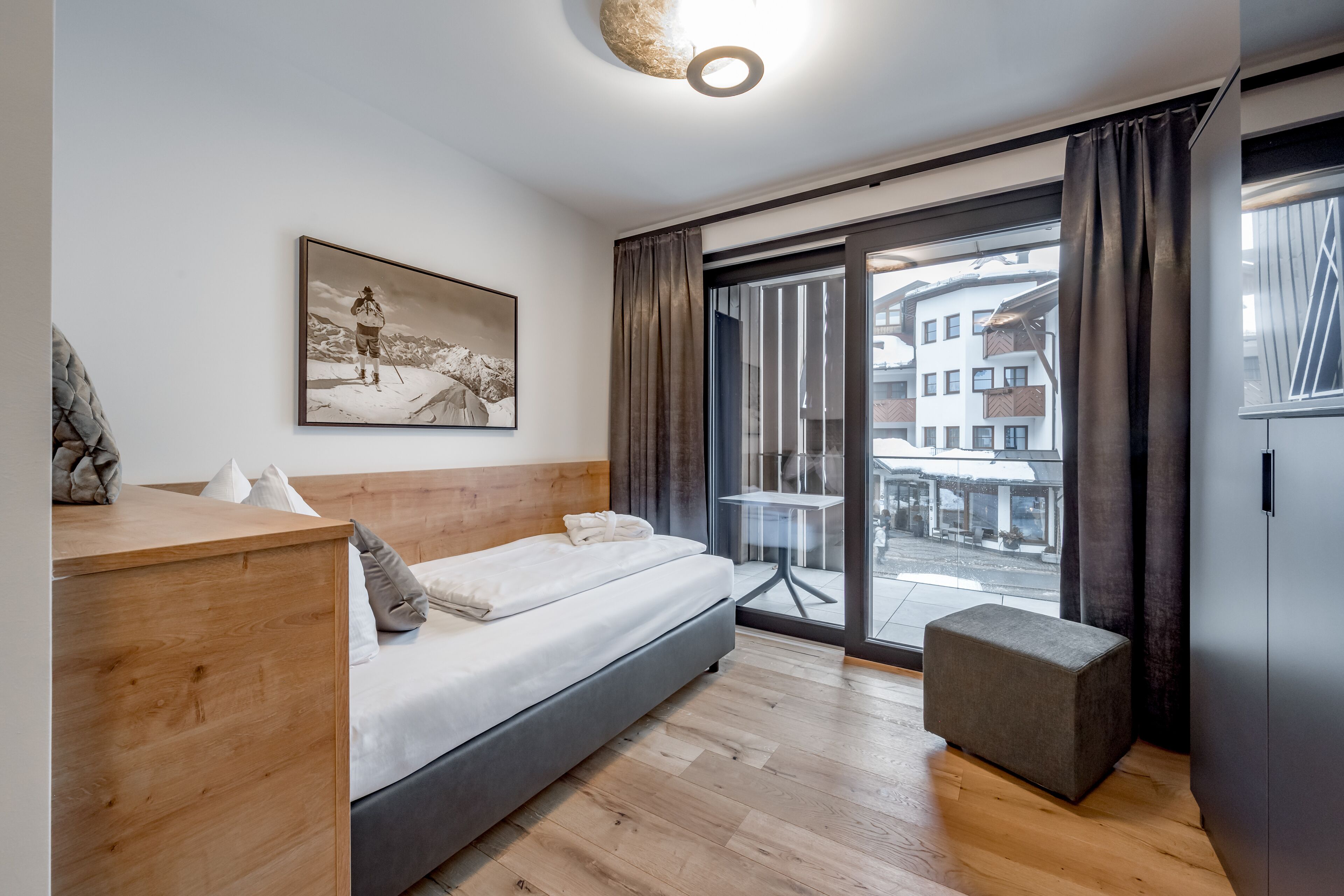 A modern bedroom with a wooden headboard, a bed with white linens, a small ottoman, a large window with curtains, and a city view.