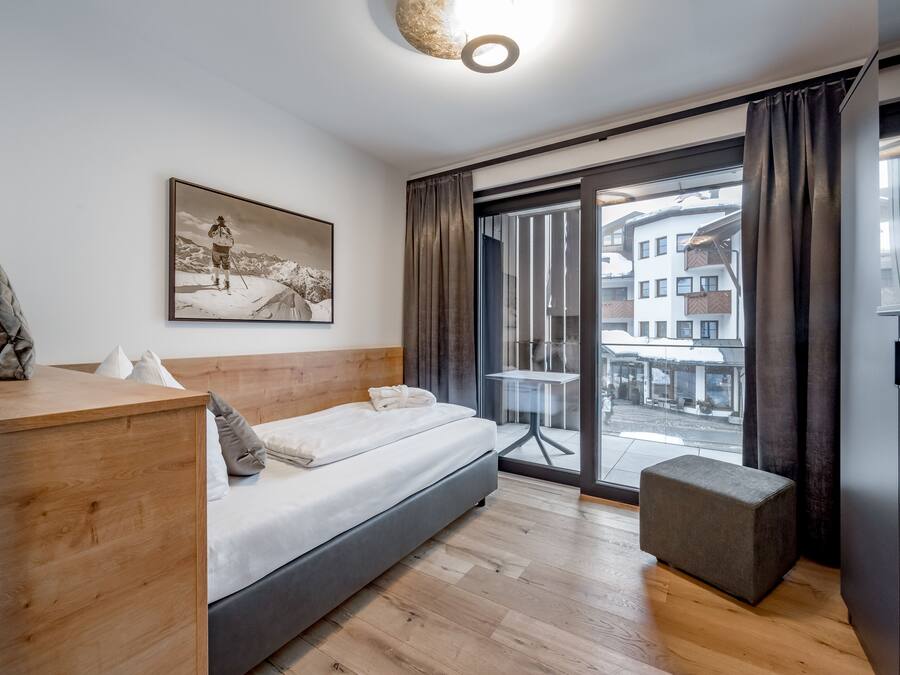 A modern bedroom with a wooden headboard, a bed with white linens, a small ottoman, a large window with curtains, and a city view.