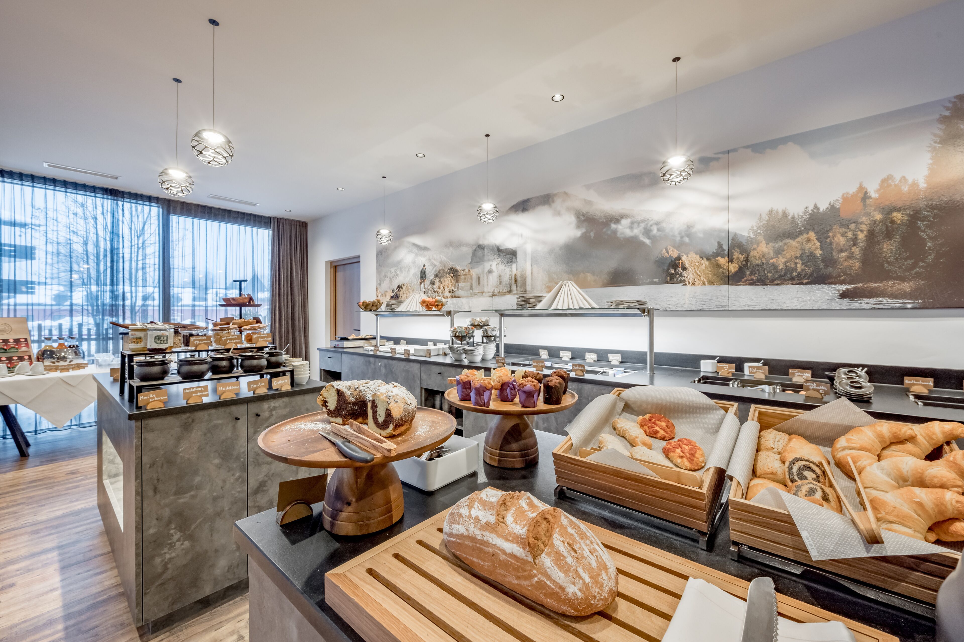 A breakfast buffet with various pastries and bread, a coffee station, and a scenic mural on the wall.