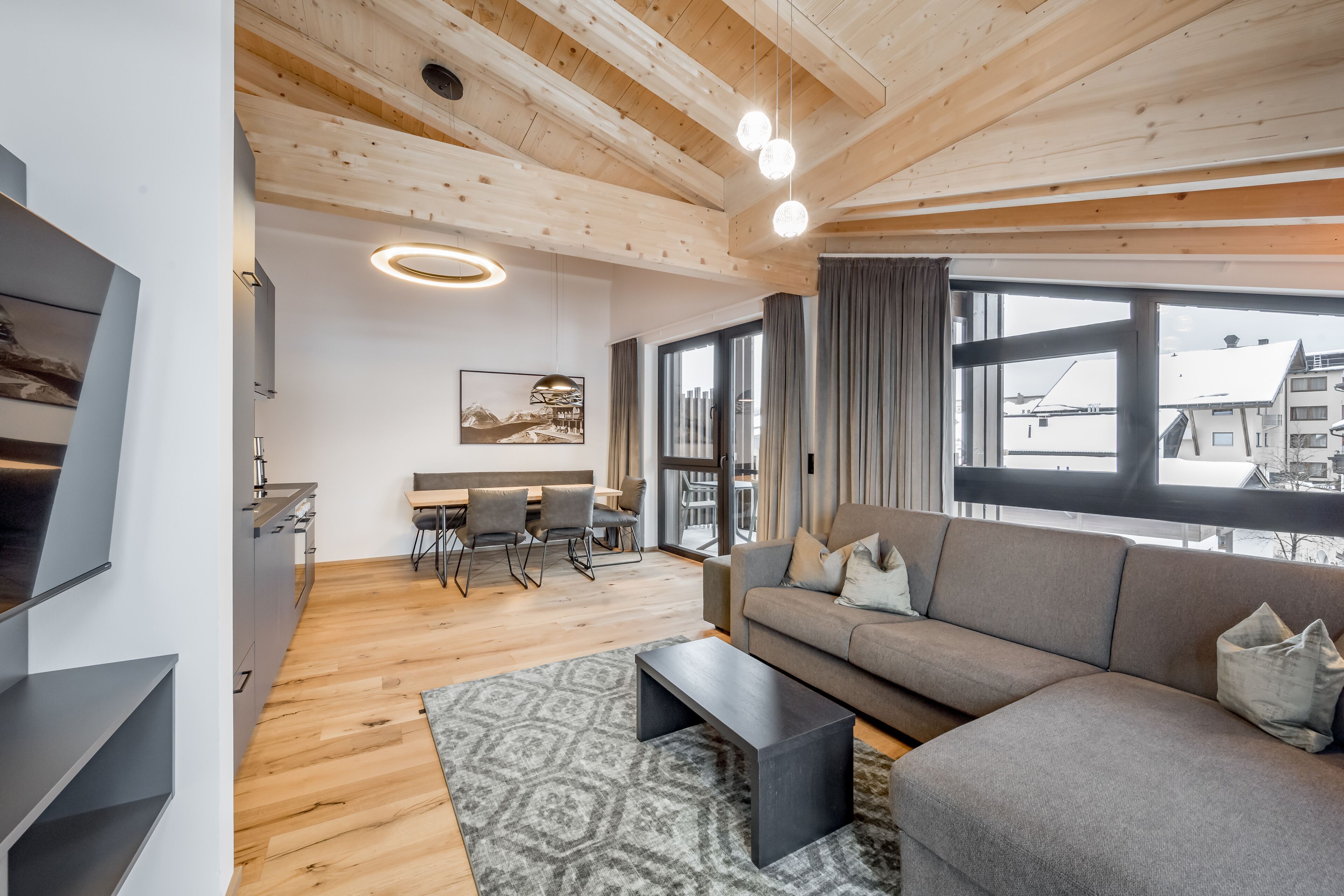 A modern living room with a sofa, coffee table, and dining area with chairs. There is a large window with curtains, a TV mounted on the wall, and wooden ceiling beams.