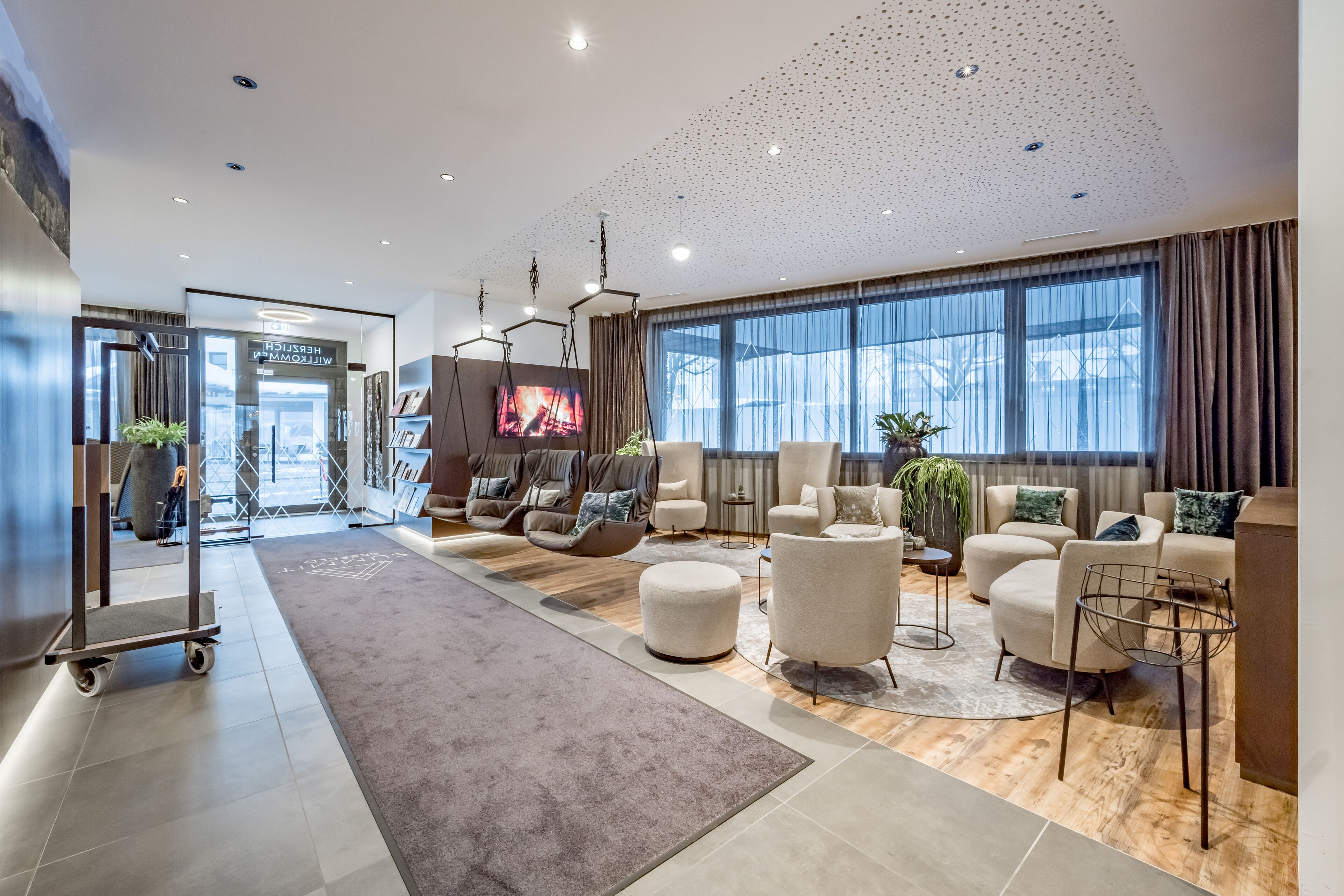 A modern hotel lobby with a spacious seating area, a large TV screen, and a view of the city through large windows.